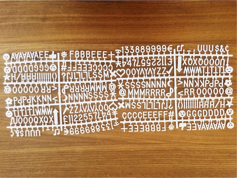 K&ouml;nnte beinhalten: Wei&szlig;e Kunststoffbuchstaben mit verschiedenen Buchstaben, Zahlen und Symbolen. Die Buchstaben sind in Reihen und Spalten angeordnet, wobei jede Kachel ein einzelnes Zeichen enth&auml;lt. Die Buchstaben befinden sich auf einer Holzoberfl&auml;che.