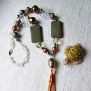 May include: A long necklace with a variety of beads, including clear, brown, and green glass beads, and two rectangular green stone pendants. The necklace has a silver tassel with an orange and white tassel. A small ceramic turtle is also pictured.