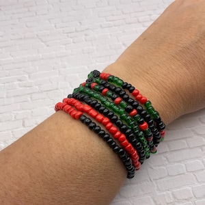 May include: A stack of beaded bracelets in red, black, and green. The bracelets are made of small, round beads and are stacked on top of each other. The bracelets are on a wrist.