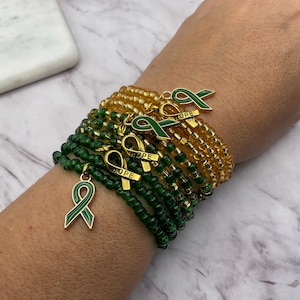 May include: A collection of beaded bracelets in shades of green and gold. Each bracelet features a gold ribbon charm with the word "HOPE" and a green enamel ribbon. The bracelets are stacked on a wrist.