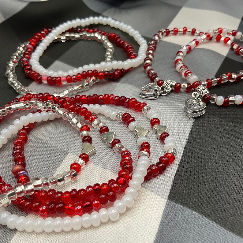 May include: Set of four red and white beaded bracelets with silver heart charms. The bracelets are layered and feature a mix of small and large beads.