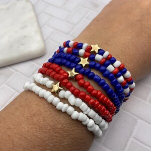 May include: A stack of beaded bracelets in red, white, and blue, with gold star charms. The bracelets are made of small, round beads and are worn on a wrist. The color scheme evokes a patriotic theme.