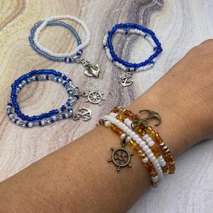 May include: Several beaded bracelets in shades of blue, white, and amber. Each bracelet features a nautical charm, including anchors and a ship's wheel. The bracelets are displayed on a person's wrist and against a neutral background.