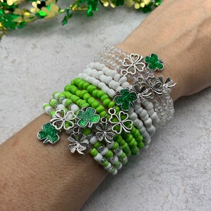 May include: A stack of green and white beaded bracelets with silver shamrock charms. The bracelets are layered on a person's wrist.