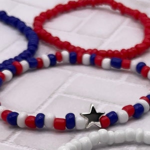 May include: Several beaded bracelets in red, white, and blue. One bracelet features a silver star charm. The bracelets are made of small, round beads, and are likely elasticated for easy wear. Patriotic theme.