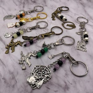 May include: A collection of seven metal keychains featuring various charms including ghosts, witches, a haunted house, and a spider web. Each keychain is accented with colorful beads in shades of black, white, green, and purple, attached to silver or bronze-toned rings and lobster clasps.