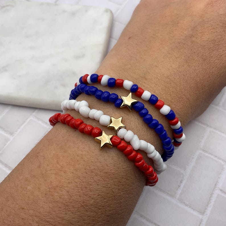 May include: Four beaded bracelets in red, white, and blue with gold star charms. The bracelets are worn on a wrist. The beads are small and round. The bracelets are a patriotic theme.
