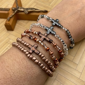 May include: A set of five bracelets with cross charms. The bracelets are made of small beads in various colors, including copper, gold, and clear. The cross charms are made of metal and have a distressed finish.