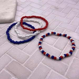 May include: Four beaded bracelets in red, white, and blue. Three bracelets are solid colors: red, white, and blue. The fourth bracelet has a pattern of red, white, and blue beads. The bracelets are on a white and gray tiled surface.