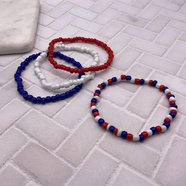 May include: Four beaded bracelets in red, white, and blue. Three bracelets are solid colors: red, white, and blue. The fourth bracelet has a pattern of red, white, and blue beads. The bracelets are on a white and gray tiled surface.