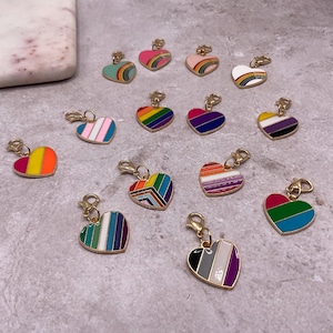 May include: A collection of heart-shaped charms with gold-colored clasps. Each charm features a different colorful design, including rainbow stripes, geometric patterns, and pride flag color schemes. The charms are displayed on a textured gray surface.