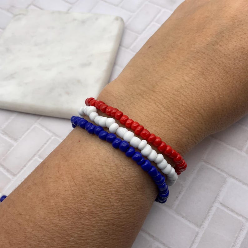 May include: Three beaded bracelets in red, white, and blue are stacked on a wrist. The bracelets are made of small, round beads. The background includes a white marble-like surface.