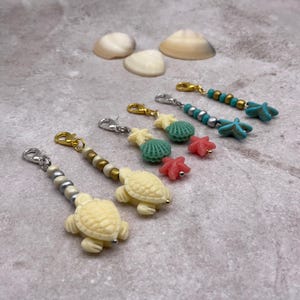 May include: A collection of beach-themed charms with lobster claw clasps. The charms feature sea turtles, starfish, and seashells in cream, turquoise, and coral colors. The charms are accented with gold and silver beads. Three seashells are in the background.