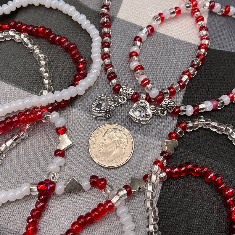May include: Five red and white beaded bracelets with silver heart charms. The bracelets are arranged on a checkered background.