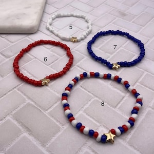 May include: Four beaded bracelets in red, white, and blue. Each bracelet features small beads and a gold star charm. The bracelets are in a patriotic color scheme, perfect for a holiday or everyday wear.