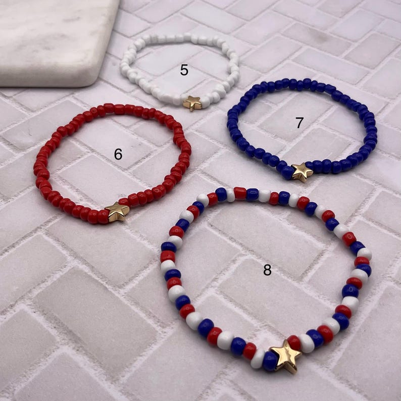 May include: Four beaded bracelets in red, white, and blue. Each bracelet features small beads and a gold star charm. The bracelets are in a patriotic color scheme, perfect for a holiday or everyday wear.