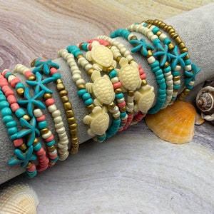 May include: Stack of beaded bracelets in shades of turquoise, coral, cream, and gold. Some bracelets feature starfish or turtle charms. The bracelets are displayed on a gray cylinder, with seashells in the background.