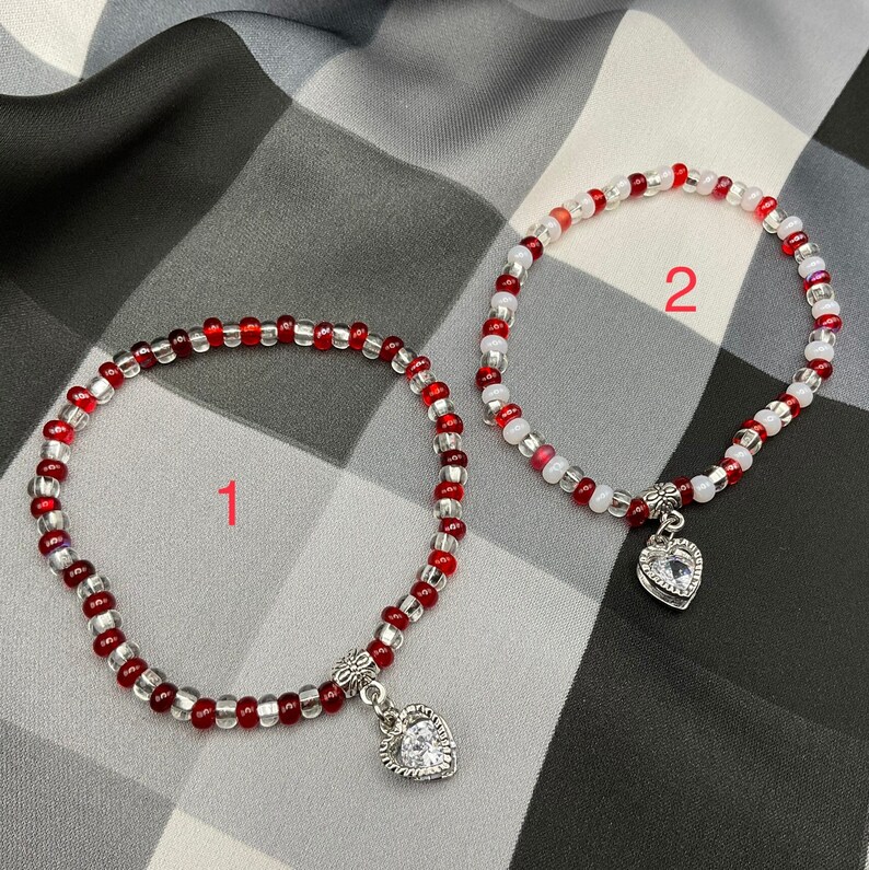 May include: Two beaded bracelets with heart charms. One bracelet is red and white, the other is white and clear. Both bracelets have a silver heart charm with a clear rhinestone.