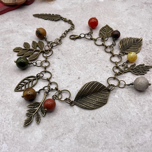 May include: A charm bracelet featuring antique bronze-toned metal chain links adorned with various leaf-shaped charms and round, multi-colored gemstone beads in shades of red, orange, yellow, green, brown, and grey, secured with a lobster claw clasp.