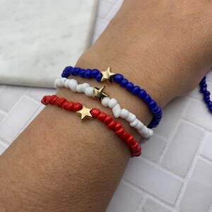 May include: Three beaded bracelets in red, white, and blue, each with a gold star charm. The bracelets are worn on a wrist, showcasing the patriotic color scheme. The beads are small and round, creating a delicate design.