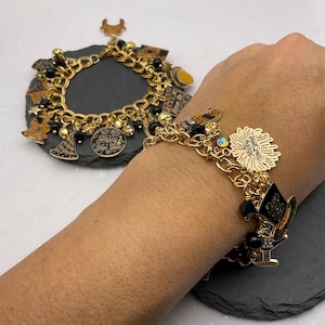May include: Gold-toned charm bracelets with black and gold charms. One bracelet is on a dark gray surface, the other is worn on a wrist. Charms include a champagne bottle, top hat, and a Happy New Year medallion.