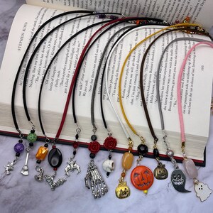 May include: A collection of Halloween-themed bookmarks with various charms, including a witch's broom, bats, a skull, and pumpkins. Each bookmark has a different colored cord and bead accents, displayed open on a book.