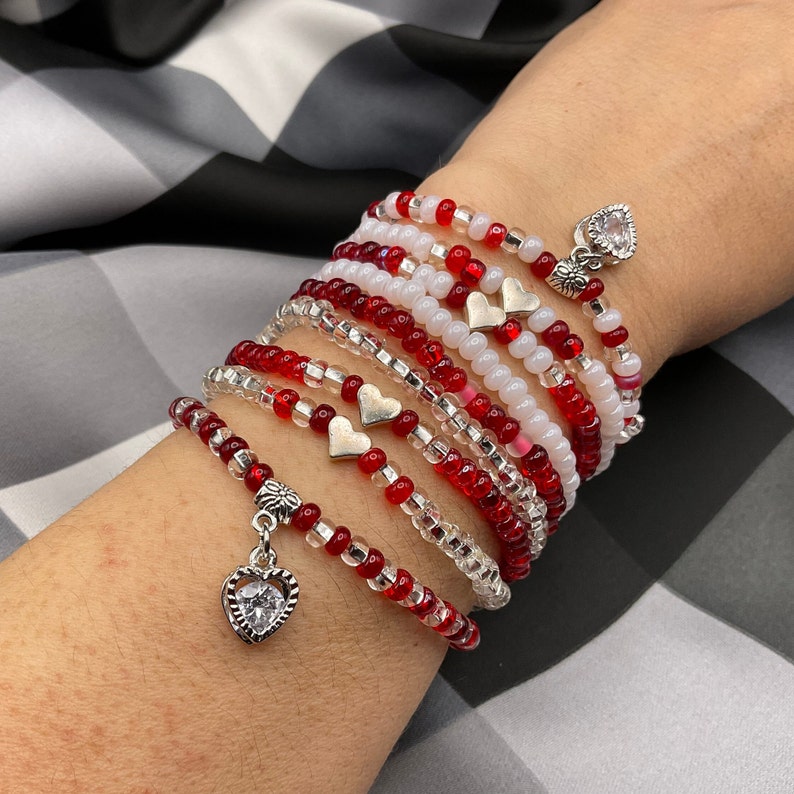 May include: A stack of red and white beaded bracelets with silver heart charms. Each bracelet has a different pattern of red and white beads.