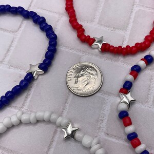 May include: Close-up of four beaded bracelets in red, white, and blue, with a silver star charm. A U.S. dime is in the center for size comparison. The bracelets are made of small, round beads, and the star charms are silver-colored.