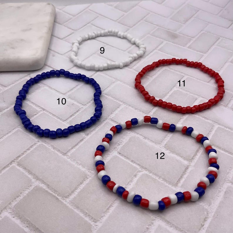 May include: Four beaded bracelets in white, blue, and red. One bracelet is white, one is blue, and one is red. The fourth bracelet has a pattern of red, white, and blue beads. The bracelets are displayed on a white and gray tiled surface.