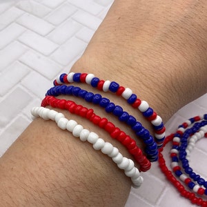 May include: A collection of beaded bracelets in red, white, and blue. The bracelets are made of small, round beads strung together. The bracelets are worn on a wrist, with additional bracelets visible in the background.