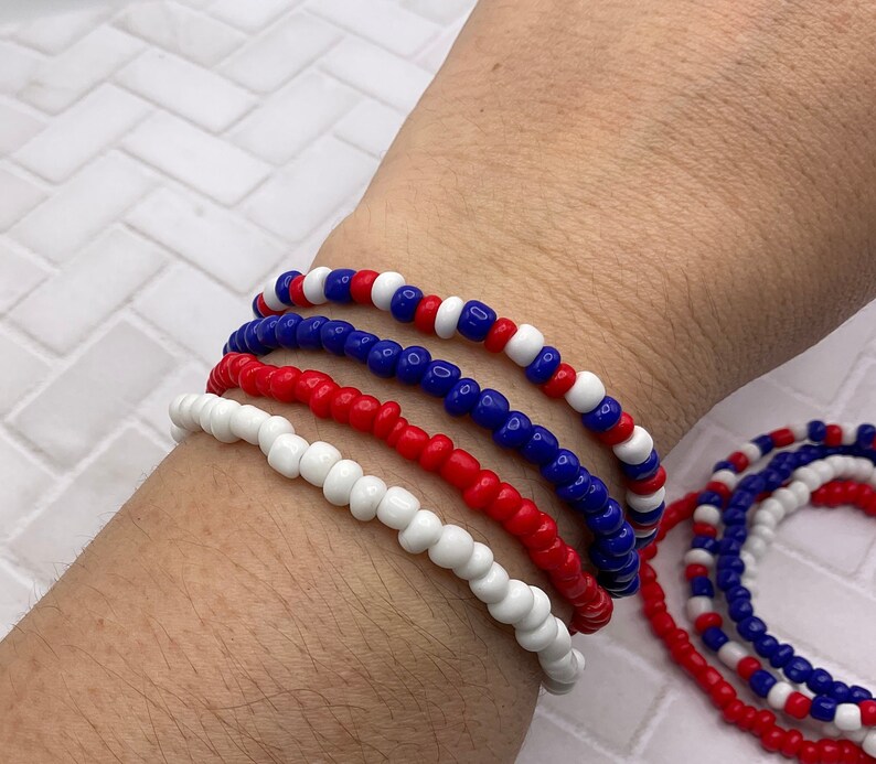 May include: A collection of beaded bracelets in red, white, and blue. The bracelets are made of small, round beads strung together. The bracelets are worn on a wrist, with additional bracelets visible in the background.