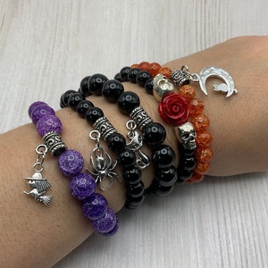 May include: Five Halloween themed bracelets with charms. The bracelets are made of black, orange, and purple beads. The charms include a skull, a cat, a spider, a rose, and a witch on a broomstick.