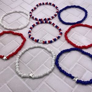 May include: A collection of beaded bracelets in red, white, and blue. Some bracelets feature a silver star charm. The bracelets are made of small, round beads and are designed to be worn on the wrist. The bracelets are a patriotic theme.
