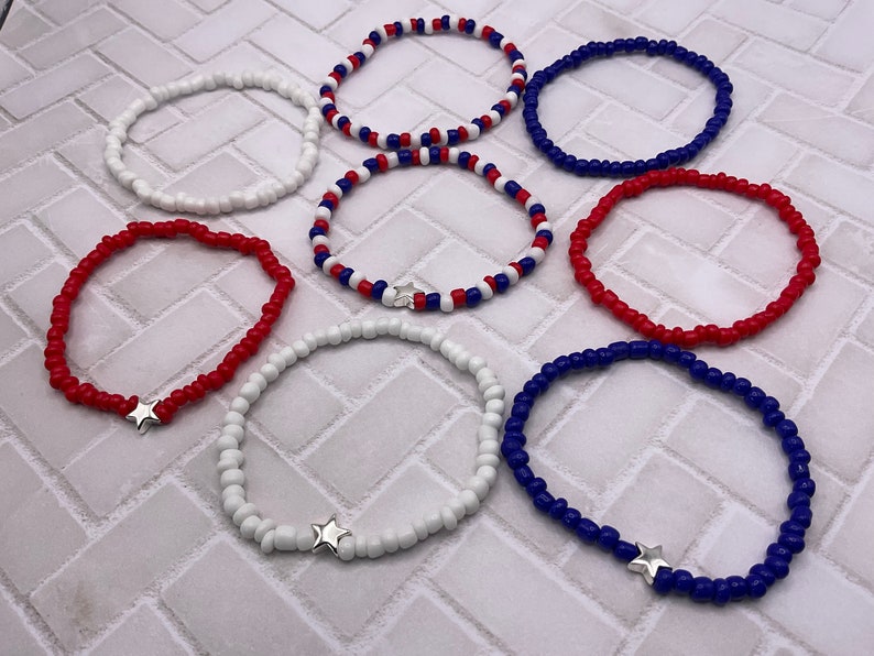 May include: A collection of beaded bracelets in red, white, and blue. Some bracelets feature a silver star charm. The bracelets are made of small, round beads and are designed to be worn on the wrist. The bracelets are a patriotic theme.