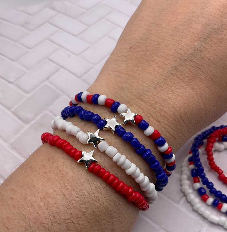 May include: Red, white, and blue beaded bracelets with silver star charms. The bracelets are worn on a wrist, showcasing the patriotic color scheme. The beads are small and evenly spaced, creating a delicate and festive accessory.