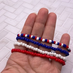 May include: Four beaded bracelets in red, white, and blue. The bracelets are made of small, round beads strung together. The colors are arranged in alternating patterns, creating a patriotic theme. The bracelets are shown against a neutral background.