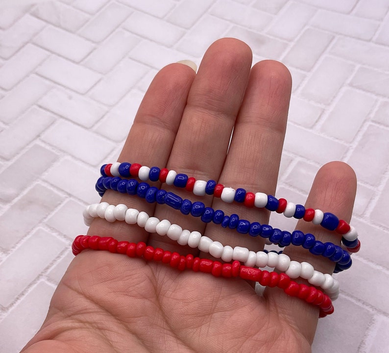 May include: Four beaded bracelets in red, white, and blue. The bracelets are made of small, round beads strung together. The colors are arranged in alternating patterns, creating a patriotic theme. The bracelets are shown against a neutral background.