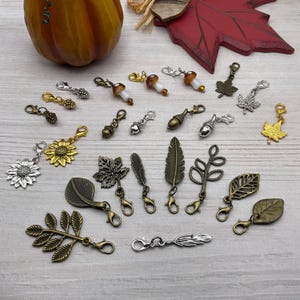 May include: A collection of decorative charms with lobster claw clasps. The charms feature various autumn-themed designs, including leaves, acorns, mushrooms, and sunflowers. The charms are in shades of gold, silver, and bronze, perfect for jewelry making or crafting.