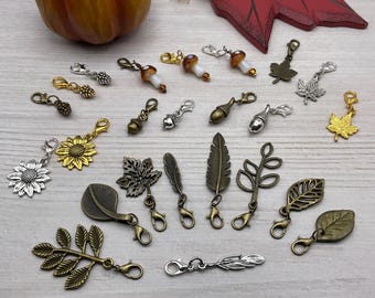 May include: A collection of decorative charms with lobster claw clasps. The charms feature various autumn-themed designs, including leaves, acorns, mushrooms, and sunflowers. The charms are in shades of gold, silver, and bronze, perfect for jewelry making or crafting.
