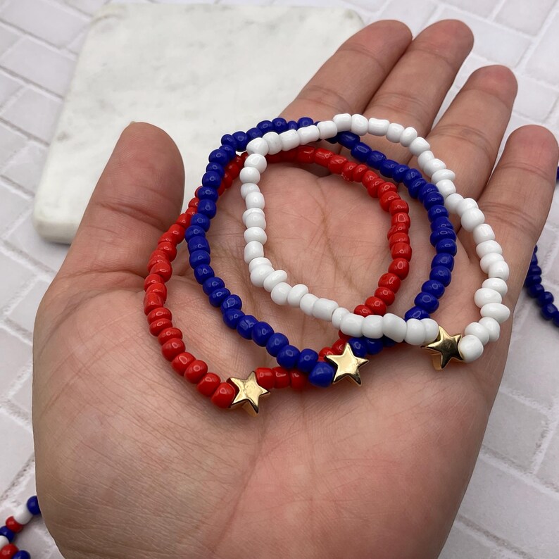 May include: Three beaded bracelets in red, white, and blue, each with a gold star charm. The bracelets are made of small, round beads and are displayed on a hand.