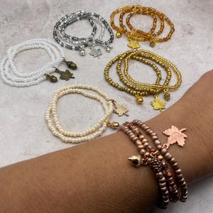 May include: A collection of multi-strand beaded bracelets featuring metallic and pearl-like finishes. Each bracelet includes small charms shaped like maple leaves, acorns, or pinecones in silver, gold, bronze, and copper tones.