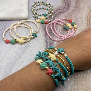 May include: Several beaded bracelets in various colors, including white, pink, and turquoise. The bracelets are adorned with sea-themed charms such as starfish, turtles, and seashells. The bracelets are displayed on a marble surface.