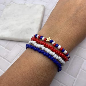 May include: Four beaded bracelets in red, white, and blue, with a gold star charm. The bracelets are stacked on a wrist, showcasing the patriotic color scheme. The beads are small and round, creating a simple yet stylish accessory.