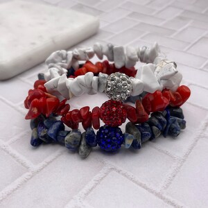 May include: Three stacked bracelets with a patriotic theme. The top bracelet features white stone chips, the middle one has red stone chips, and the bottom one has blue stone chips. Each bracelet has a decorative bead with rhinestones.