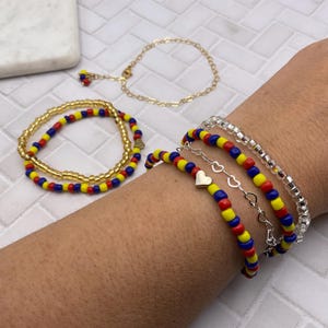 May include: Several beaded bracelets in red, yellow, and blue are displayed. One bracelet is gold with a heart charm. Another is a gold chain bracelet. A clear beaded bracelet is also shown. The bracelets are on a wrist.