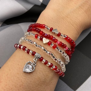 May include: A set of five red and silver beaded bracelets with heart-shaped charms. The bracelets are stacked on a person's wrist.