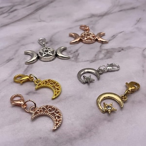 May include: Five silver, gold, and rose gold charm pendants with a moon and star design, a triple moon and pentagram design, and a crescent moon design. The charms are on a white marble background.