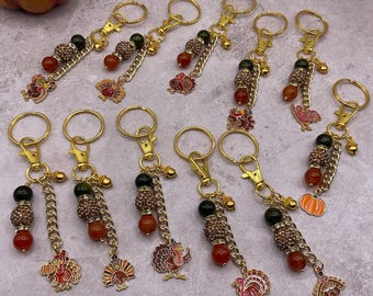 May include: A collection of gold-tone metal keychains featuring turkey and pumpkin charms, accented with brown, orange, and black beads, small gold bells, and chain links. Each keychain includes a lobster clasp for attachment.