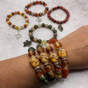 May include: A collection of beaded bracelets featuring various natural stone and glass beads in shades of amber, green, red, and yellow. Several bracelets include silver-toned metal charms shaped like leaves and butterflies, with some featuring rhinestone spacers.