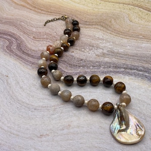 May include: A necklace featuring alternating round faceted beads in shades of brown, tan, and gray, accented by small white spacer beads. The necklace includes a large, iridescent, teardrop-shaped shell pendant and a bronze-toned metal chain clasp.